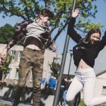 Two teenagers joyfully jumping outdoors in Šiluva, Lithuania under a bright blue sky. Captured in springtime.